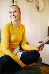 Porträt einer glücklichen blonden Frau mit digitalem Tablet, die auf einem Sessel zu Hause sitzt und in die Ferne schaut - DAWF01473