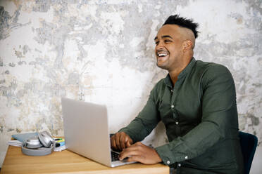 Porträt eines lachenden jungen Mannes, der im Büro zu Hause am Laptop arbeitet - DAWF01475