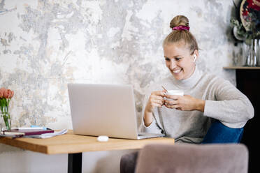 Smiling blond woman sitting on armchair at home having video chat with laptop and earphones - DAWF01481