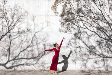 Woman in red dress with dancer pose and tree shadows on wall - TCEF00624