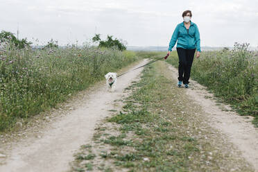 Ältere Frau mit Schutzmaske geht mit ihrem Hund auf einem Feldweg spazieren - XLGF00147