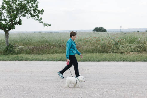 Rückenansicht einer älteren Frau, die mit ihrem Hund auf dem Land spazieren geht - XLGF00148