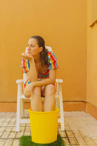 Portrait of serious woman relaxing on plastic chair cooling her legs in bucket of water stock photo
