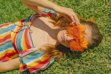 Portrait of woman lying on a meadow wearing glasses with orange flowers covering her eyes - ERRF03688