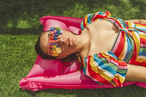 Portrait of woman wearing glasses with colourful pom poms covering her eyes relaxing on pink airbed - ERRF03695
