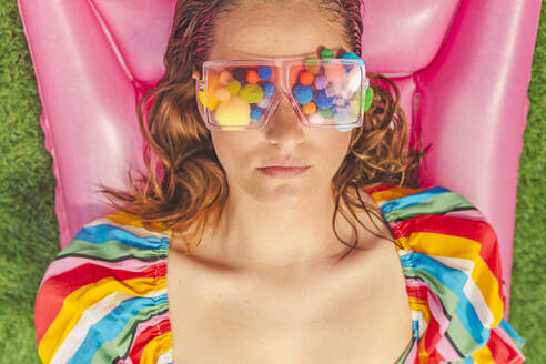 Portrait of woman wearing glasses with colourful pom poms covering her eyes relaxing on pink airbed - ERRF03696