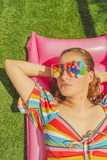 Portrait of woman wearing glasses with colourful pom poms covering her eyes relaxing on pink airbed - ERRF03697