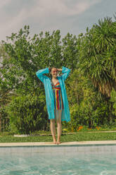 Smiling woman wearing blue rain coat standing at poolside - ERRF03714