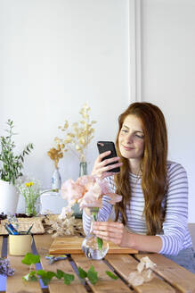 Spanien, Porträt einer jungen, schönen, rothaarigen Frau, die ihr Smartphone benutzt, während sie am Tisch Pflanzen eintopft - AFVF06246