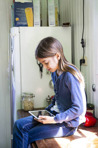 Mädchen sitzt auf der Arbeitsplatte in der Küche und schaut auf ein digitales Tablet, lizenzfreies Stockfoto