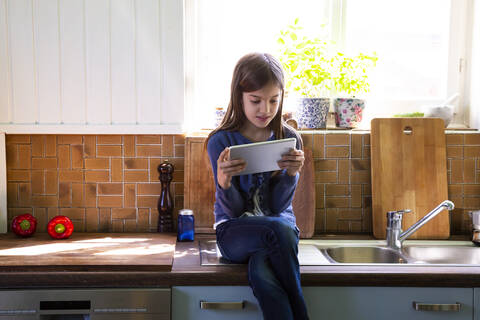 Mädchen sitzt am Waschbecken in der Küche und schaut auf ein digitales Tablet, lizenzfreies Stockfoto