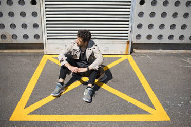 Hilfloser Mann sitzt auf dem Boden im gelb markierten Bereich vor einer Betonmauer - MAUF03407