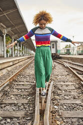 Full length of young woman with afro hairstyle walking on tracks at station against sky - VEGF02169