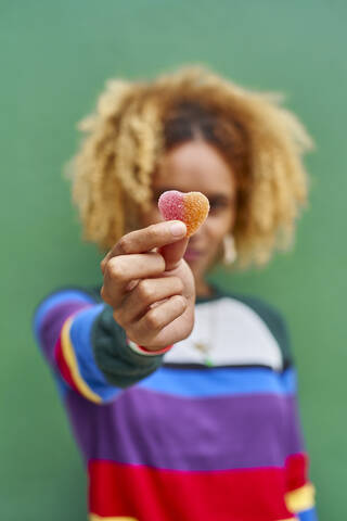 Young woman holding candy heart while standing against green wall stock photo