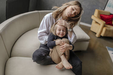Blonde Frau umarmt ihren Sohn mit der Hand auf der Stirn, während sie zu Hause auf dem Sofa sitzt - OGF00398