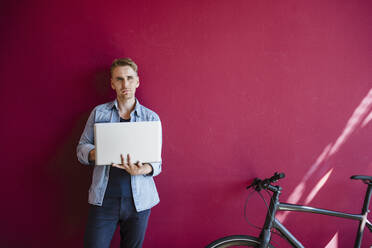 Pensive businessman leaning against red wall, using laptop - DIGF10830