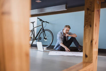 Lässiger Geschäftsmann sitzt auf dem Boden eines Loft-Büros und arbeitet an einem Bauplan - DIGF10833