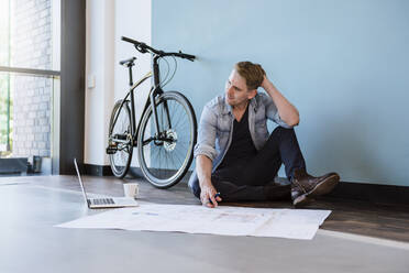 Lässiger Geschäftsmann sitzt auf dem Boden eines Loft-Büros und arbeitet an einem Bauplan - DIGF10834