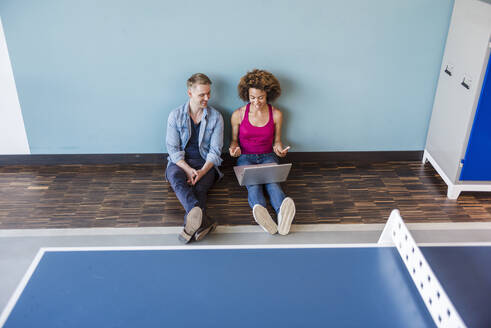 Junge Frau und Mann sitzen in einem modernen Büro auf dem Boden und benutzen einen Laptop - DIGF10857