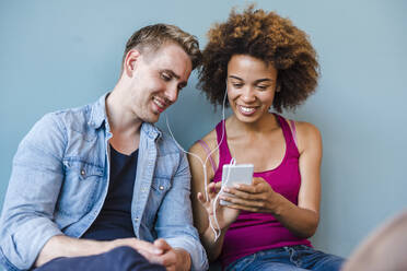 Junge Frau und Mann sitzen in einem modernen Büro auf dem Boden, teilen sich Kopfhörer und benutzen ein Smartphone - DIGF10862