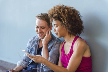 Young woman and man sitting on floor in modern office, sharing earphones, using smartphone - DIGF10863
