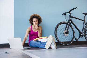 Young woman sitting on floor, eating pizza, using smartphone and earphones - DIGF10874