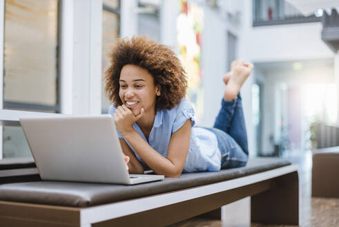 Junge Frau liegt auf einer Bank in einem modernen Büro und benutzt einen Laptop - DIGF10889