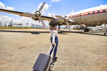 Rückansicht einer jungen Frau, die ihr Gepäck auf dem Weg zum Flugzeug am Flughafen an einem sonnigen Tag zieht - VEGF02180