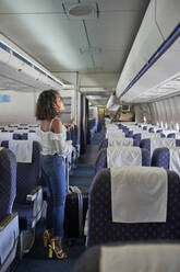 Junger weiblicher Passagier auf der Suche nach einem Sitzplatz in voller Länge, während er im Gang eines Flugzeugs steht - VEGF02184
