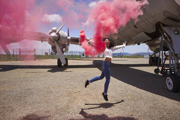 Carefree young woman enjoying while holding distress flare with pink smoke against airplane at airport runway - VEGF02189