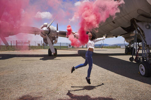 Unbeschwerte junge Frau genießt, während sie eine Notfackel mit rosa Rauch gegen ein Flugzeug auf der Startbahn eines Flughafens hält - VEGF02189