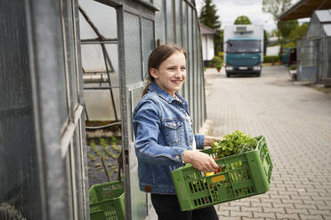 Mädchen trägt eine Kiste mit frischem Gemüse in einer Gärtnerei mit einem Lieferwagen im Hintergrund - AUF00468