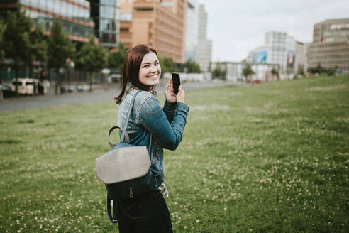 Deutschland, Berlin, Porträt einer jungen Frau, die mit einem Smartphone in der Hand in die Kamera lächelt - VBF00039