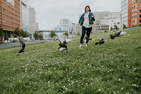 Deutschland, Berlin, Junge Frau läuft auf Gras und überrascht Taubenschwarm - VBF00040