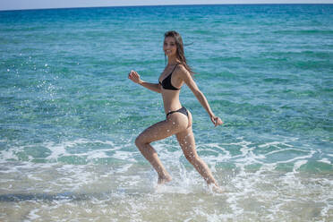 Young woman wearing bikini running in shallow water on sandy beach, smiling at camera. - CUF54967