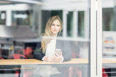 Smiling blond woman holding smartphone sitting at a window in a cafe in Berlin, Germany. - CUF55001