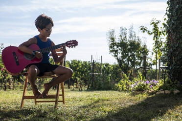 A boy sitting on a chair in the garden playing a guitar. - CUF55004