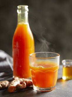 Eine Flasche und ein Trinkglas mit leuchtend orangefarbenem Wintertee neben einer Kurkumawurzel. - CUF55013