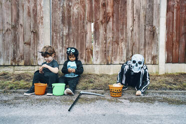 Three children dressed for Halloween, one as a skeleton, sitting at the side of a road with their buckets of sweets. - CUF55031