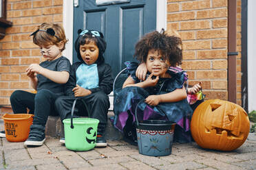 Drei für Halloween verkleidete Kinder sitzen auf einer Türschwelle und essen Süßigkeiten. - CUF55049