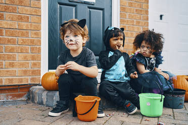 Drei zu Halloween verkleidete Kinder sitzen auf einer Türschwelle und essen Süßigkeiten. - CUF55054