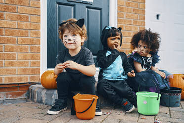 Three children dressed up for Halloween sitting on a doorstep eating sweets. - CUF55054