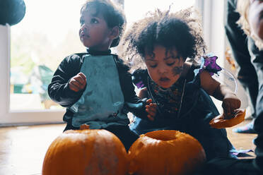 Zwei kleine Kinder, die sich für Halloween verkleidet haben und mit zwei Kürbissen auf dem Boden knien. - CUF55061