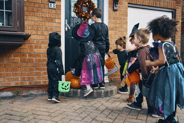 Eine Gruppe von Kindern, die sich für Halloween verkleidet haben, steht mit Eimern vor einer Haustür und gibt Süßes oder Saures. - CUF55062