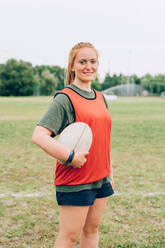 Eine Frau in Shorts und T-Shirt steht auf einem Trainingsplatz und hält einen Rugbyball. - CUF55081