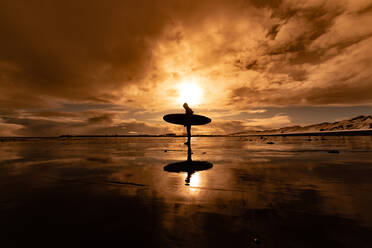 Silhouette einer Frau mit einem Surfbrett, die über einen Strand läuft, mit einem Sonnenuntergang im Hintergrund. - ISF24044