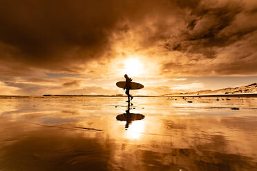 Silhouette einer Frau mit einem Surfbrett, die über einen Strand läuft, mit einem Sonnenuntergang im Hintergrund. - ISF24050