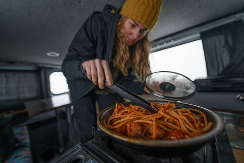 Eine Frau in einem Wohnmobil, die mit einer Wollmütze Essen in einem Kochtopf umrührt - ein winterlicher Road Trip. - ISF24051