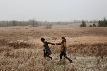 Eine schwarze Frau und ein weißer Mann halten sich an den Händen und gehen durch Gräser in einer winterlichen ländlichen Landschaft. - ISF24066