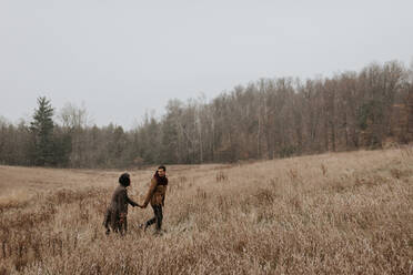 Ein Paar, das sich an den Händen hält und sich anschaut, während es durch lange Gräser in einer winterlichen Landschaft geht. - ISF24069